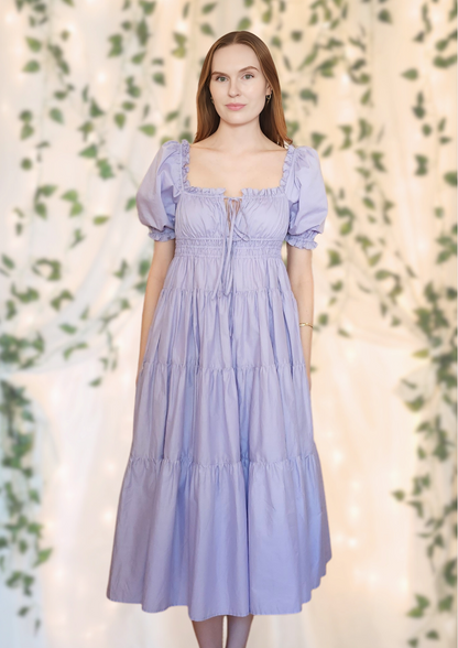 Elegant lilac midi dress with puff sleeves – the Penelope Dress by Eravela, crafted from organic cotton with a tiered skirt, adjustable bow ties, and a flattering cinched waist. Perfect for weddings, brunch, garden parties, and summer occasions. A feminine and timeless flowy dress designed for effortless elegance and comfort.